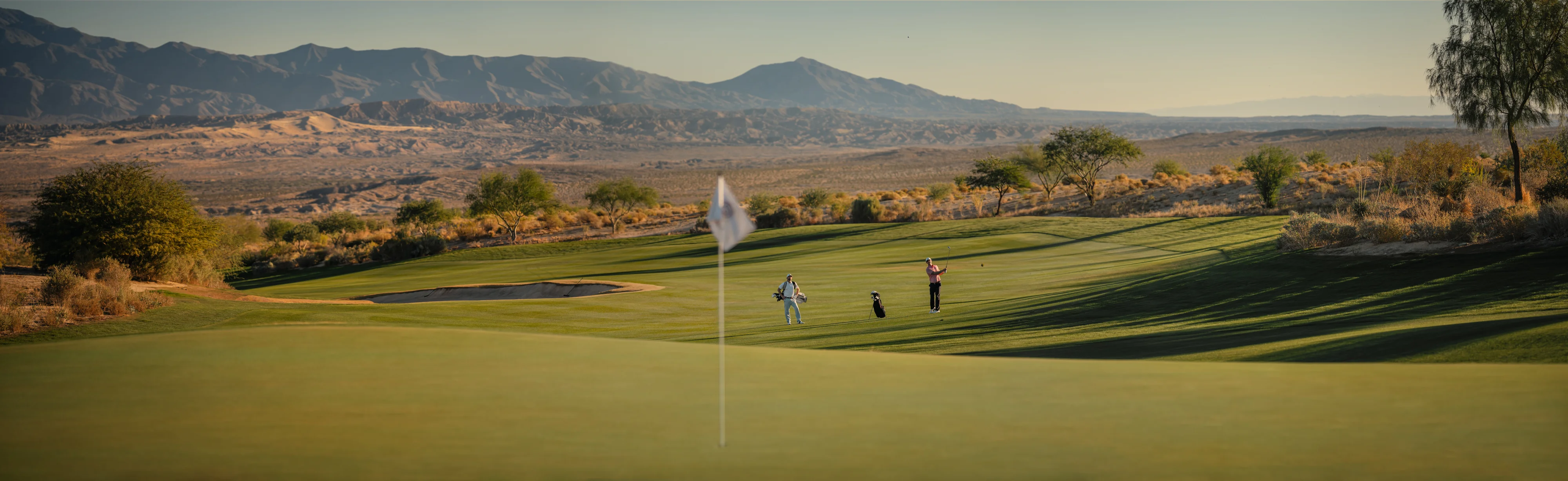 A golfer swings to hit the ball on a golf course aiming towards the hole, while another golfer observes. The backdrop features scenic mountains.