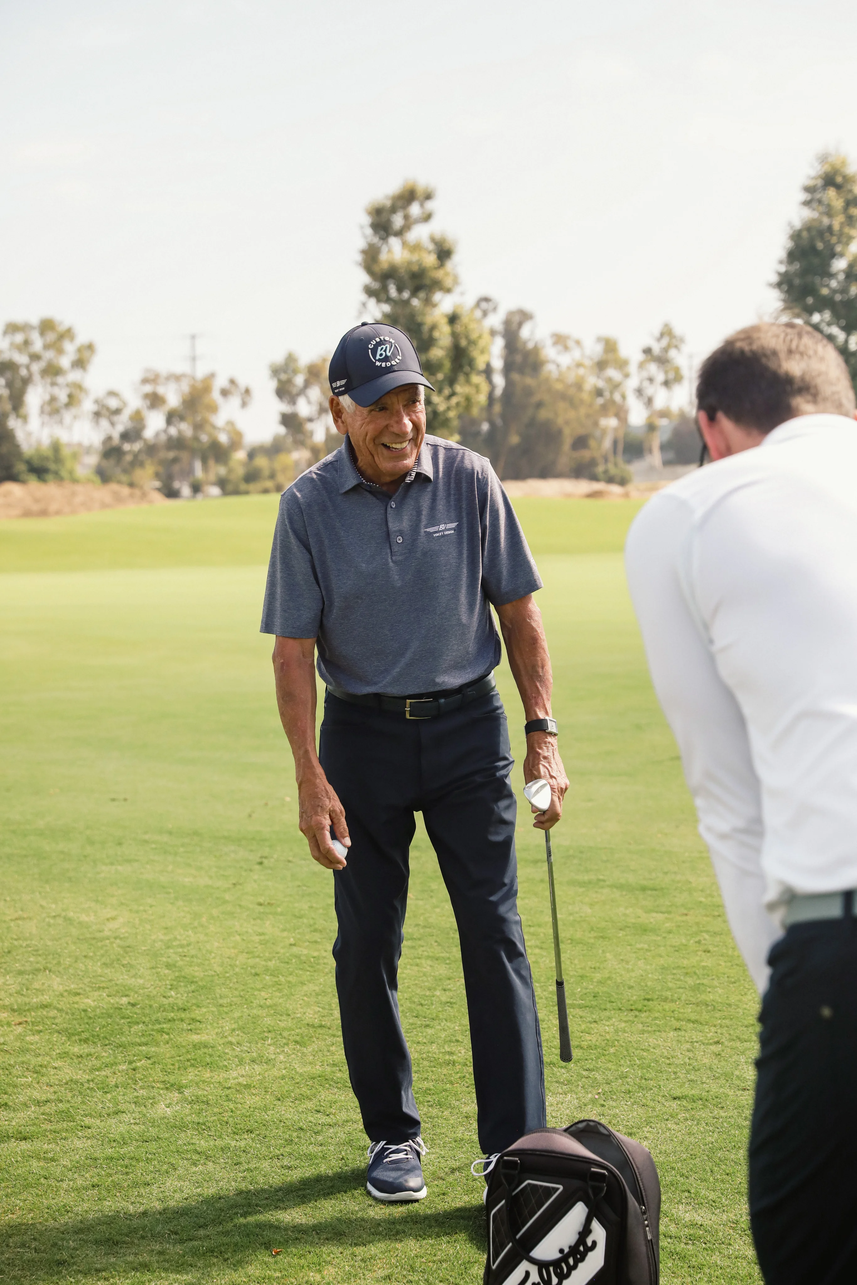Bob Vokey smiles as he holds an iron club and golf ball on the course.